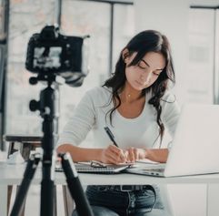 young woman working on a computer writing notes with a camera in front of her