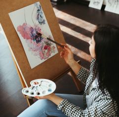 an adult woman sitting at an easel with a watercolor painting in front of her