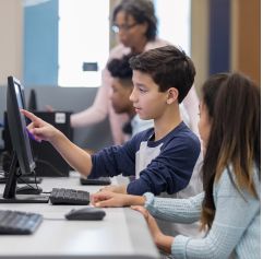 two kids sitting in front of computers with an adult in the background assisting them